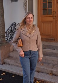 Blazer à carreaux brun clair avec de larges revers, associé à un jean bleu. Tient un petit sac à main orange. Présente une silhouette ajustée.