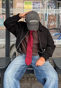 Crna bomber jakna sa srebrnim patentnim zatvaračima, smeđa košulja, crvena kravata i izblijedjele plave traperice. Siva kapa s vezom "BROOKLYN 1947".