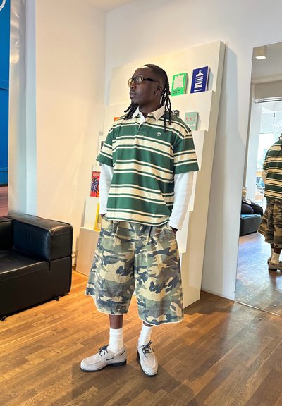 Hombre con gafas y rastas que lleva un polo de rayas de manga larga, pantalones cortos de camuflaje y zapatos beige, de pie en una habitación con libros y espejo.