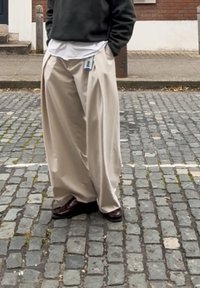 Pantaloni bej, cu croială largă, cu buzunare laterale și etichetă. Purtați peste o cămașă albă și asortați cu pantofi maro închis. Fundal cu stradă pavată.
