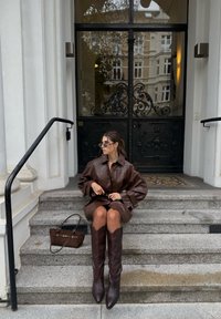 Bruine leren jas met brede mouwen, taille-ingesnoerd, en bruin met hoge hakken. Zonnebril en een bruine tas complementeren de outfit.