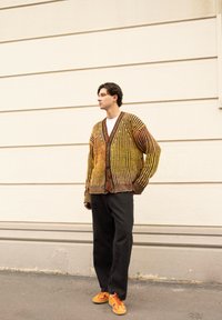 Ριγέ κα cardigan σε κίτρινο, πορτοκαλί και καφέ πλέξιμο, με V-λαιμό και κουμπί, συνδυασμένο με μαύρο φαρδύ παντελόνι και πορτοκαλί αθλητ�ικά παπούτσια.