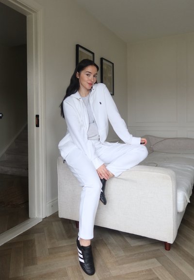 Sudadera blanca con cremallera, camiseta gris debajo, pantalones jogger blancos y zapatillas negras con rayas blancas. Sentado en un sofá de tela clara.