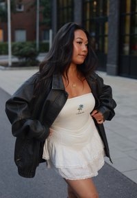 Čierna kožená oversize bunda cez bielu vrstvenú šaty s grafickým motívom modrej ruže. Oblečenie dopĺňa jemný zlatý náhrdelník.