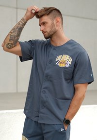 Jersey a rete grigia con bottoni, con logo "Los Angeles Lakers" in giallo e blu. Accompagnato da pantaloni corti abbinati; abbigliamento sportivo casual.