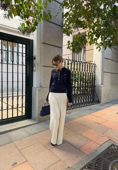 Mujer con gafas de sol, suéter azul marino con botones y pantalones blancos de pierna ancha, sosteniendo un bolso azul marino, de pie en una acera de baldosas bajo ramas frondosas.