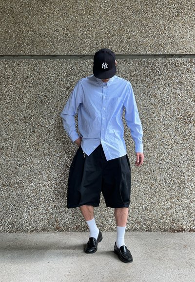 Camisa de botones a rayas azul y blanca, pantalones cortos negros oversize, calcetines blancos y mocasines negros, bajo una gorra negra, de pie sobre una superficie texturizada.