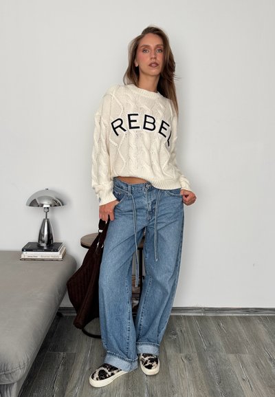 Suéter blanco de punto con el texto "REBEL", jeans azules holgados, zapatos de plataforma con estampado y un bolso marrón; entorno interior con una lámpara y libros.