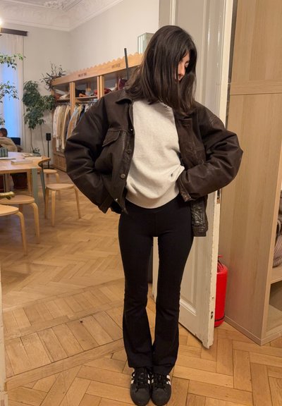 Mujer joven con pantalones negros, suéter blanco y chaqueta marrón, de pie en el interior de una habitación con suelos de madera y percheros de ropa.