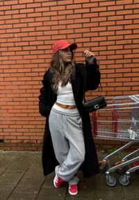Černý dlouhý kabát, bílý crop top, šedé tepláky a červené tenisky. Červená čepice, černá kabelka na rameno. Kovové nákupní vozíky poblíž na pozadí cihlové zdi.
