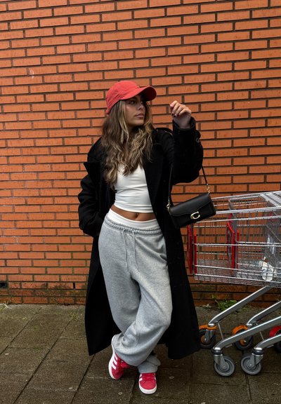 Abrigo largo negro, top corto blanco, pantalones deportivos grises y zapatillas rojas. Gorra roja, bolso negro cruzado. Carritos de compras metálicos cerca, junto a una pared de ladrillos.