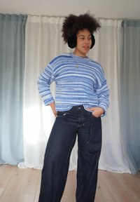 Blå og hvit stripet strikkegenser med lange ermer, kombinert med mørke denimbukser med vide ben. Modellen har på seg svarte ørevarmere.