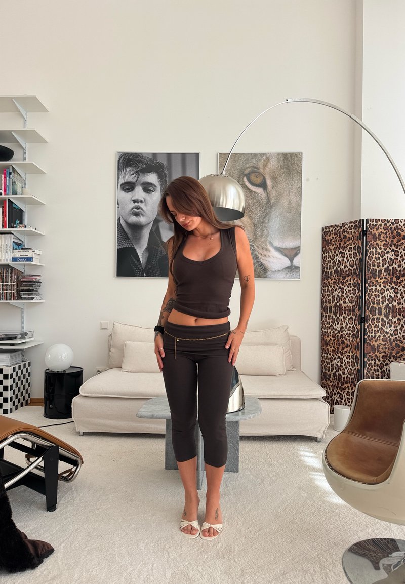 Femme en haut noir sans manches et leggings debout dans un salon moderne avec un canapé beige, un paravent à motif léopard et de grands portraits de lion et d’homme.
