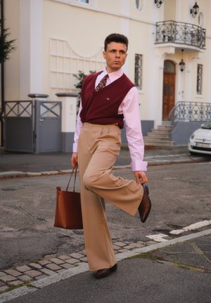 Bordeauxfarbener Weste über einem pinken Hemd, gemusterte Krawatte, beiger weit geschnittener Hose, braunen Lederschuhen und einer braunen Tragetasche, auf einer Straße platziert.