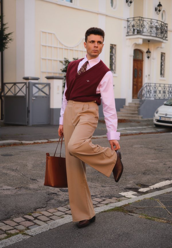Gilet bordeaux su una camicia rosa, cravatta con motivo, pantaloni beige a gamba larga, scarpe in pelle marroni e una borsa a spalla marrone, posizionati su una strada.