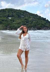 Copricostume da spiaggia bianco trasparente con maniche lunghe e coulisse in vita, decorato con un accento floreale. Modello in piedi a piedi nudi sulla sabbia bagnata vicino al mare.