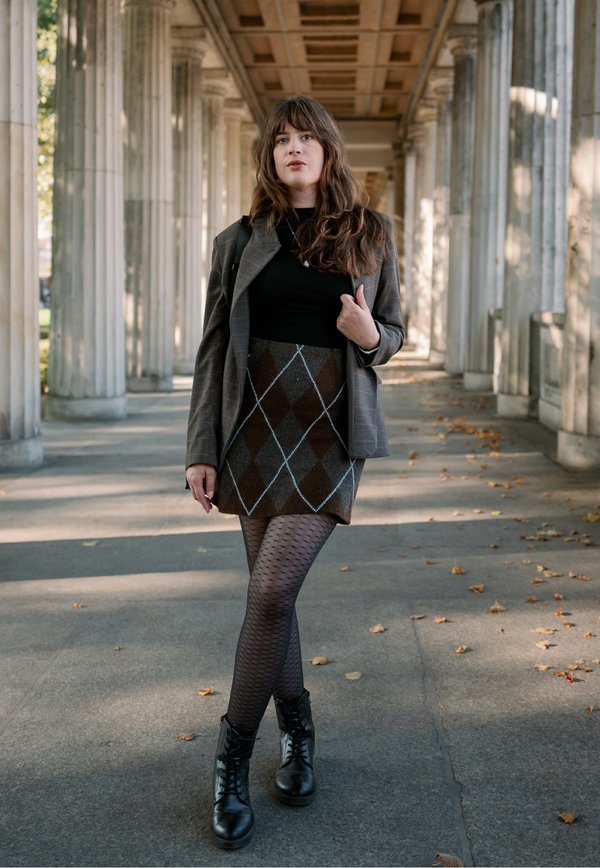 Une femme en blazer gris, jupe à motifs, collants et bottes marche sous une colonnade en tenant une bretelle de sac à dos.