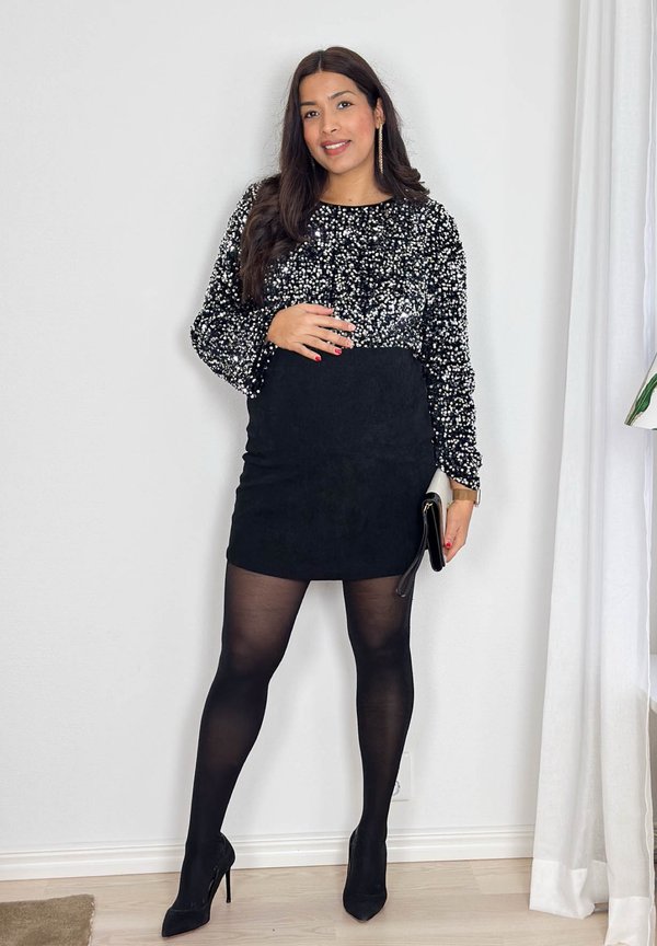 Vrouw poseert binnenshuis, gekleed in een zwarte top met pailletten, een zwarte minirok, zwarte doorzichtige panty en zwarte hoge hakken, terwijl ze een kleine clutch vasthoudt.