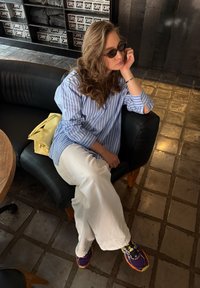 Striped blue and white shirt, white wide-leg trousers, purple and yellow trainers. Sitting on a black leather sofa with a yellow bag.