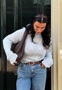 Lys grå kabelstrik sweater, blå højtaljede jeans, brun fletet taske og matchende bælte. Sort hår med en brun pandebånd.