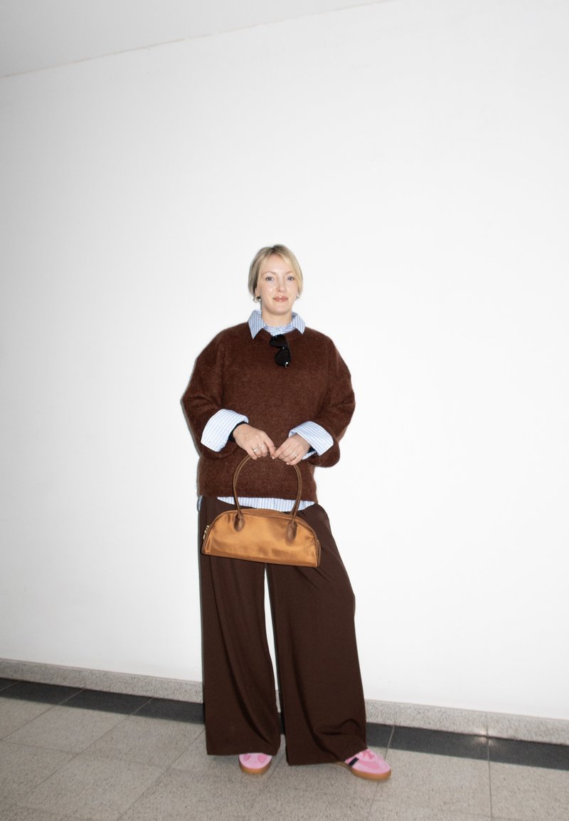 Femme en pull marron et pantalon large tenant un sac à main beige, portant des chaussures roses, debout contre un mur blanc.