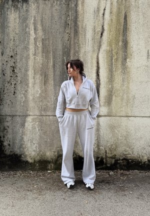 Mujer con sudadera con capucha gris claro recortada con cremallera y pantalones deportivos a juego, de pie con las manos en los bolsillos frente a una pared de concreto envejecida.