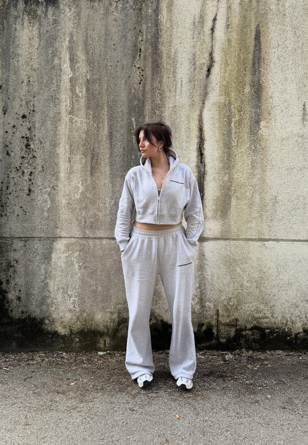 Γυναίκα με ανοιχτό γκρι cropped φούτερ με φερμουάρ και ταιριαστό παντελόνι φόρμας στέκεται με τα χέρια στις τσέπες μπροστά σε φθαρμένο τσιμεντένιο τοίχο.
