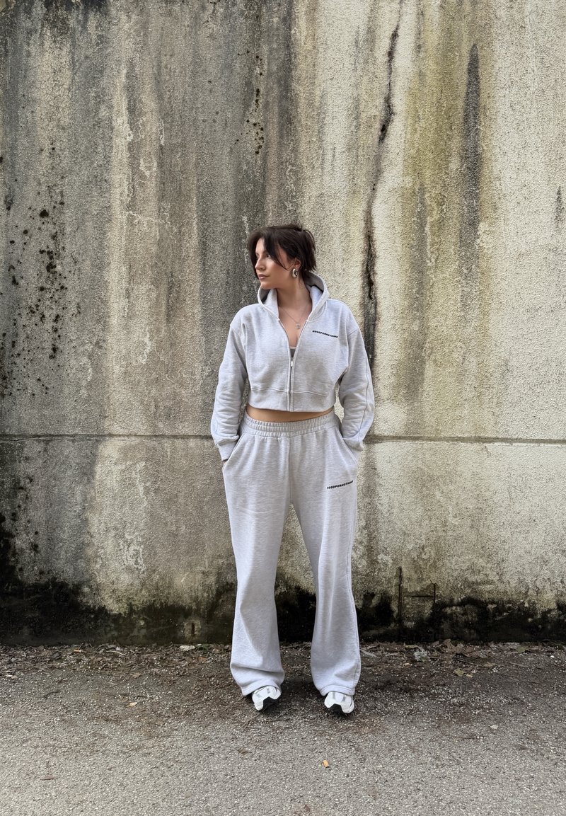 Femme portant un sweat à capuche gris clair court zippé et un pantalon de survêtement assorti, debout les mains dans les poches contre un mur de béton usé.