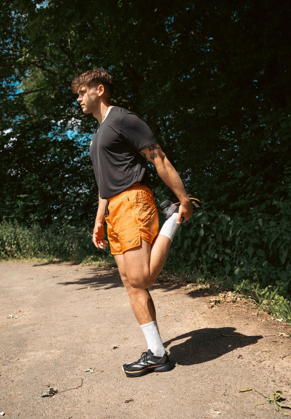 Athlète s'étirant sur un chemin, portant un t-shirt noir à manches courtes, un short orange, des chaussettes blanches et des chaussures de course noires. Feuillage vert en arrière-plan.