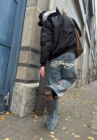 Zwarte bomberjack, grijze hoodie, blauwe spijkerbroek met wit "Ed Hardy" logo en kleurrijke geborduurde patches, zwarte schoenen op kinderkopjes.