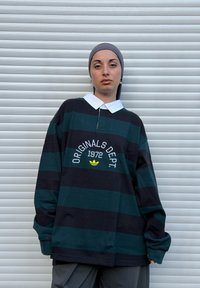 Personne portant un foulard gris, une chemise ample à rayures noires et vert foncé avec le texte « ORIGINALS DEPT. 1972 », debout devant un volet métallique blanc.