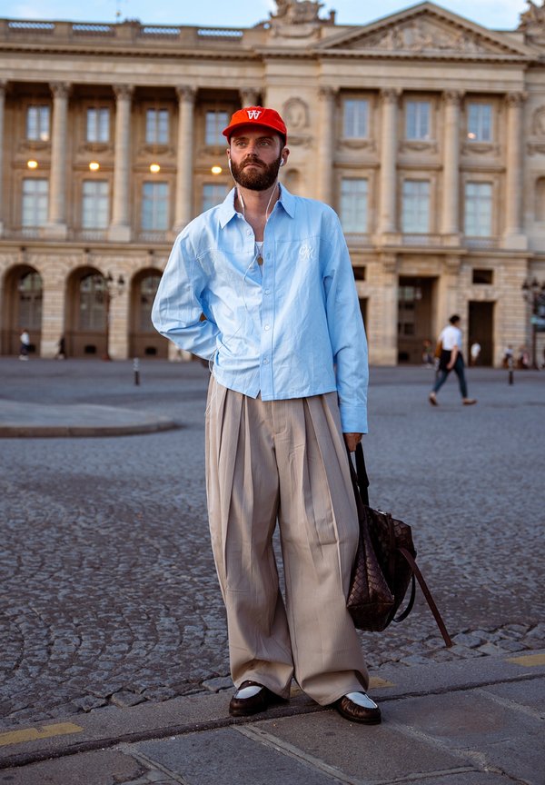 Man met baard, die een rode pet, lichtblauw hemd, wijde beige broek en witte schoenen draagt, staat op een geplaveide straat en houdt een bruine tas vast.