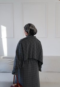 Femme portant un manteau à carreaux foncé et un foulard à motifs, tenant un sac à main marron, debout dans une pièce lumineuse et minimaliste avec un canapé beige.