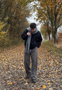 Juoda striukė, pilkas šalikas, pilkos kelnės. Rudens lapija supa žmogų, einantį taku, išklotu oranžiniais ir geltonais lapais.