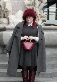 Femme portant une robe grise, un col roulé blanc, un manteau gris, un chapeau en fourrure rouge, tenant un sac à main rouge, debout sur des marches en pierre devant une fontaine sculptée.