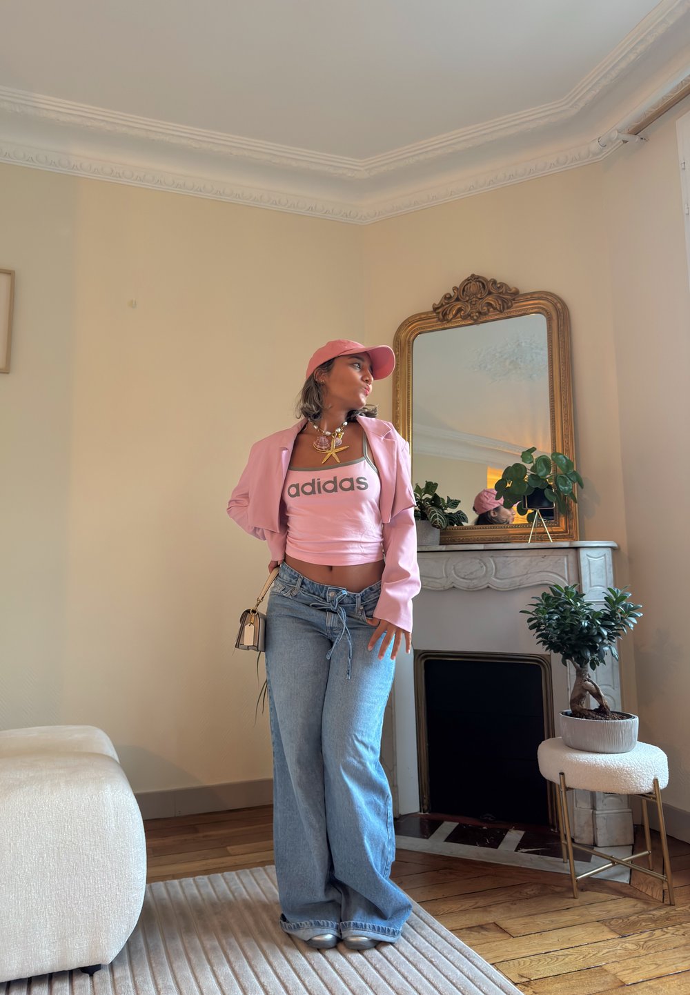 Giovane donna in top Adidas rosa, con cappellino e giacca abbinati, jeans blu si posa in un soggiorno con pavimento in legno, camino e piante d'appartamento.