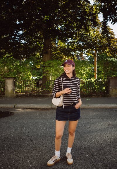 Camisa de rayas de manga larga, falda de mezclilla oscura, zapatillas blancas y una gorra oscura; sosteniendo un vaso para llevar; bolso de hombro; fondo urbano.