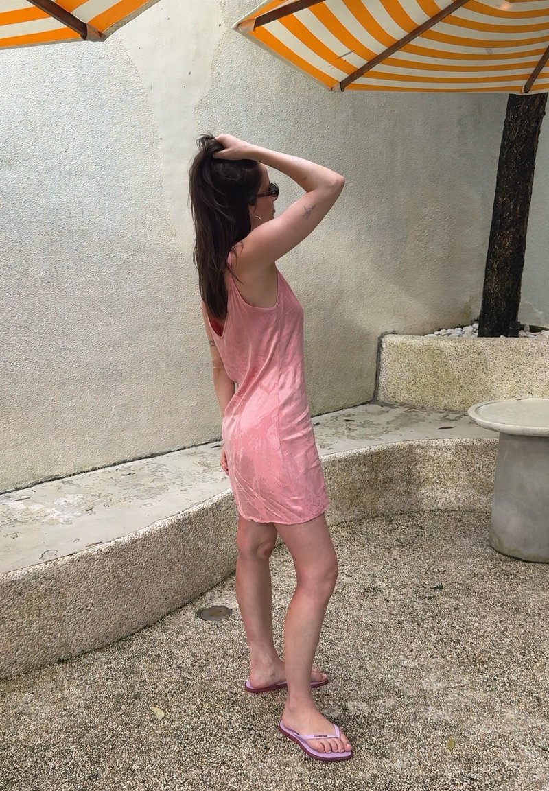 Kvinde i pink ærmeløs kjole og klip-klapper står under stribede gule og hvide parasoller og rører ved sit hår med den ene hånd.