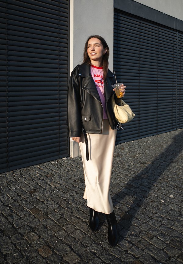 Giovane donna che indossa giacca nera in pelle, T-shirt rosa con grafica, gonna color crema, tiene in mano una bevanda e una borsa beige, in piedi su una strada acciottolata.