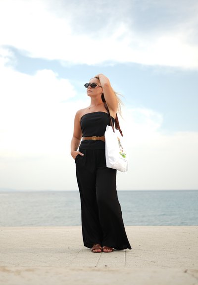 Mono negro sin tirantes con un cinturón, sandalias de beige arenoso y una bolsa tote blanca. Pelo largo y gafas de sol, de pie junto a la orilla del agua.