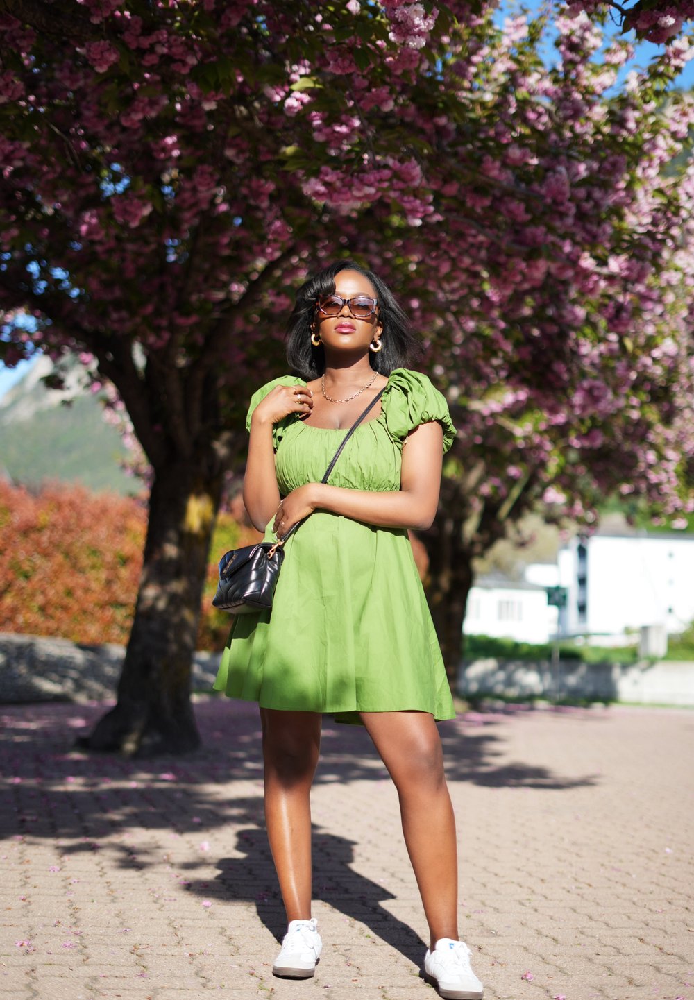Robe courte verte avec des manches bouffantes, en tissu texturé. Portée avec des baskets blanches et un sac à main noir. Entourée d'arbres en fleurs.