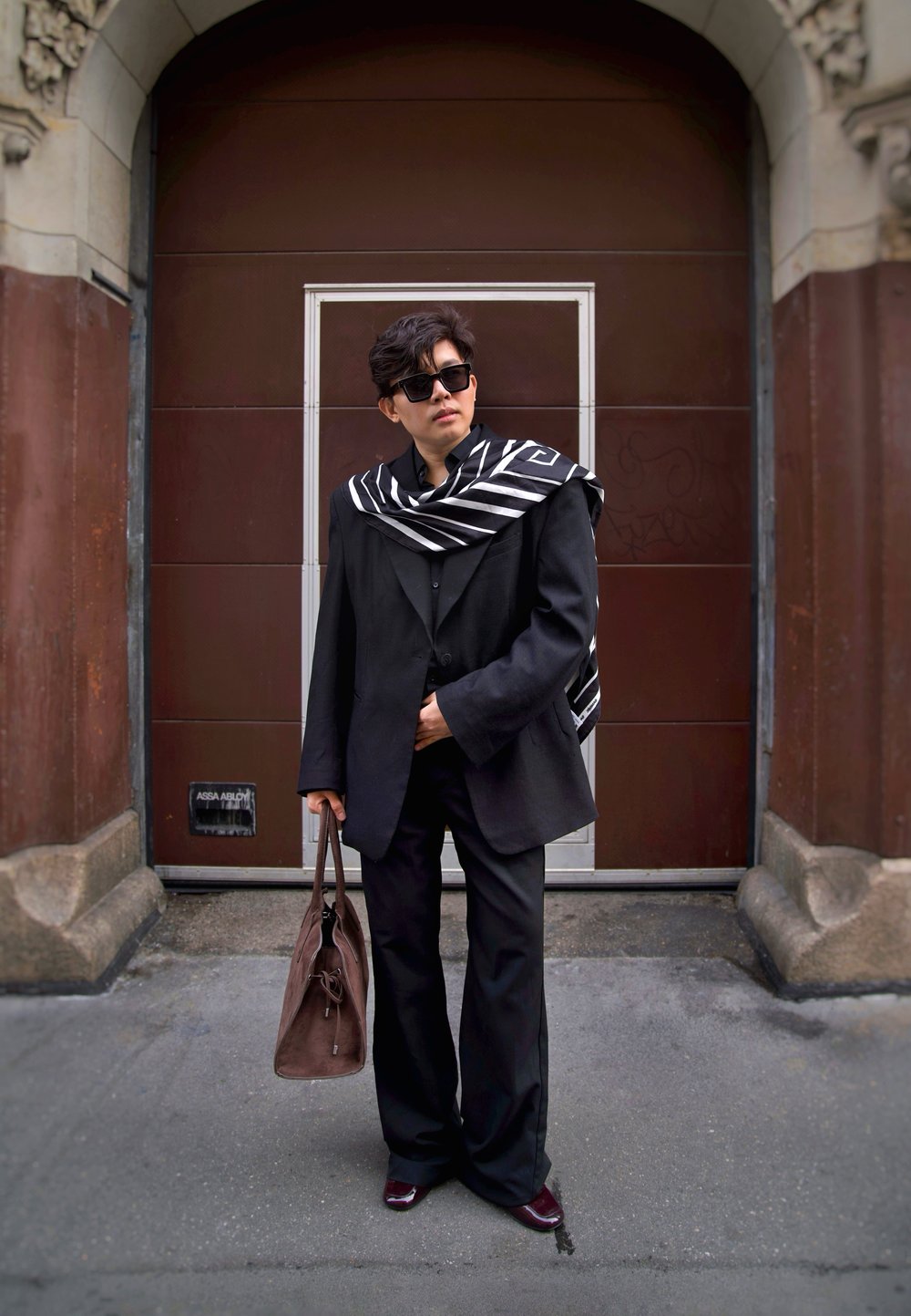 Crni oversized sako s pripadajućim hlačama, slojevit s crno-bijelim prugastim šalom. Izgled zaokružuju smeđa torba i tamne sunčane naočale.