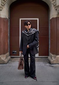Crni oversized sako s pripadajućim hlačama, slojevit s crno-bijelim prugastim šalom. Izgled zaokružuju smeđa torba i tamne sunčane naočale.
