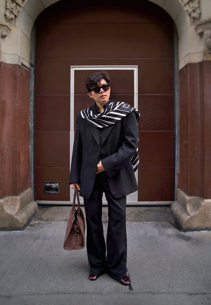 Crni oversized sako s pripadajućim hlačama, slojevit s crno-bijelim prugastim šalom. Izgled zaokružuju smeđa torba i tamne sunčane naočale.