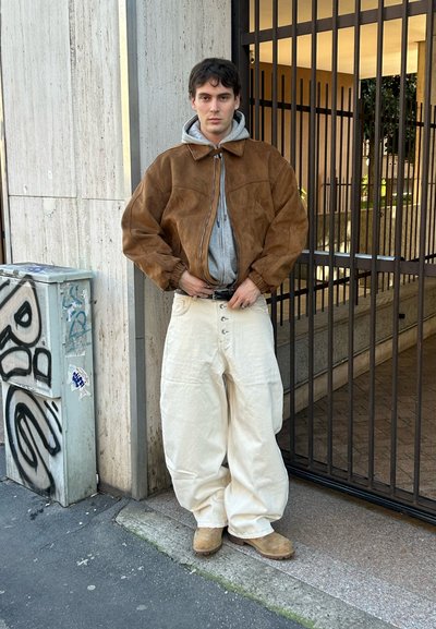 Joven con chaqueta marrón de ante, sudadera gris con capucha, pantalones crema holgados y botas color canela, de pie en una acera de la ciudad cerca de una entrada con reja.