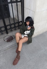 Grüne gesteppte Jacke mit braunem Kragen, weiß-grün gestreiftem Pullover, braunen Shorts, strukturierten braunen Socken und braunen Slippern.