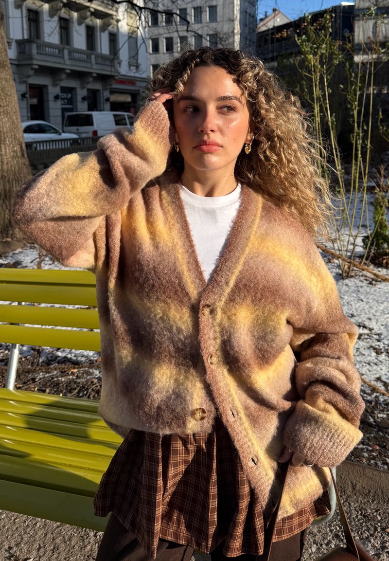 Jeune femme aux cheveux bouclés portant un cardigan multicolore en matière duveteuse et une jupe à carreaux marron, debout près d'un banc jaune dans un parc urbain ensoleillé.