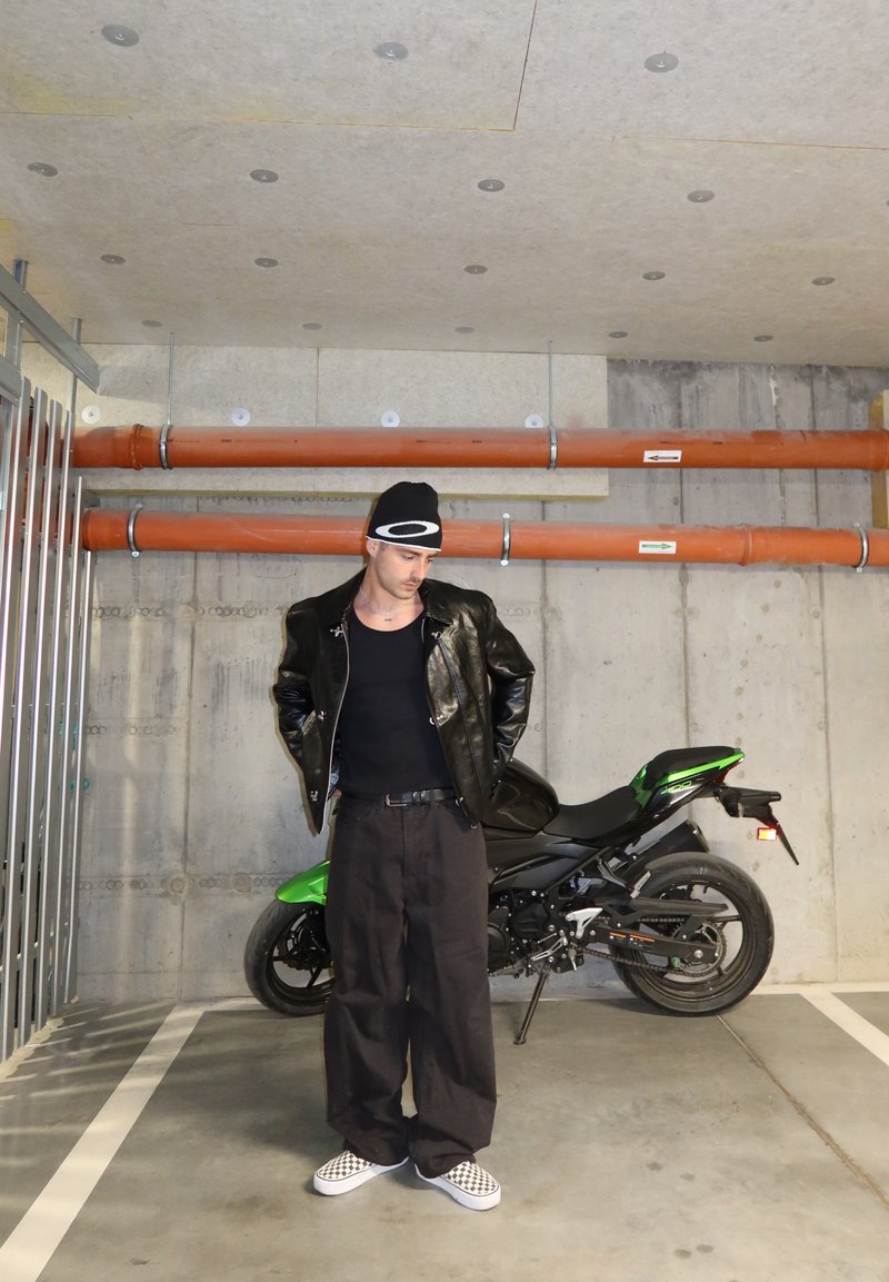 Mann in schwarzer Lederjacke, schwarzem Tanktop, Mütze und karierten Schuhen steht in einer Betongarage neben einem grünen und schwarzen Motorrad.