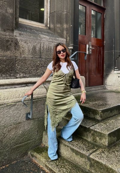Mujer con gafas de sol, camiseta blanca, vestido verde oliva y jeans azules posando en escalones de piedra frente a un edificio con puertas de madera.