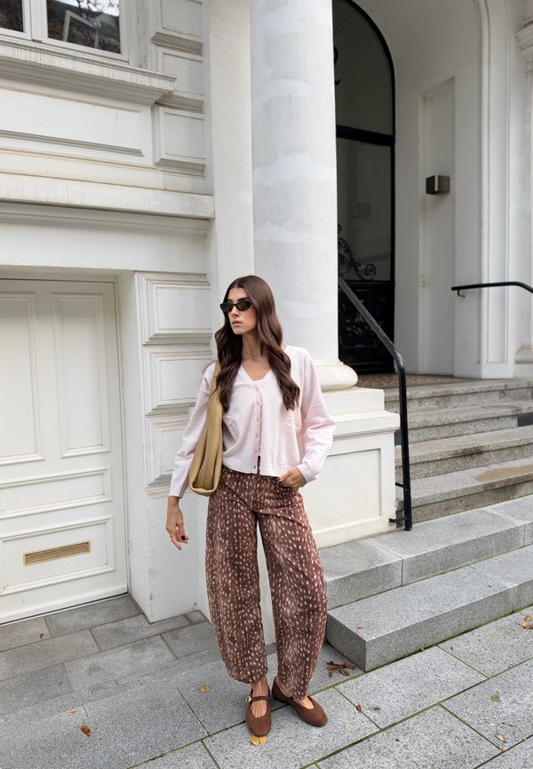 Une femme portant des lunettes de soleil, un cardigan rose pâle, un pantalon à motifs et des chaussures plates se tient à côté de marches en pierre, devant un bâtiment blanc.